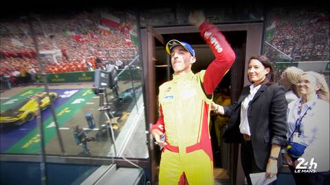 Kierowca wyścigowy w ż&oacute;łto-czerwonym kombinezonie świętuje, wznosząc rękę, na tle trybun i toru wyścigowego.