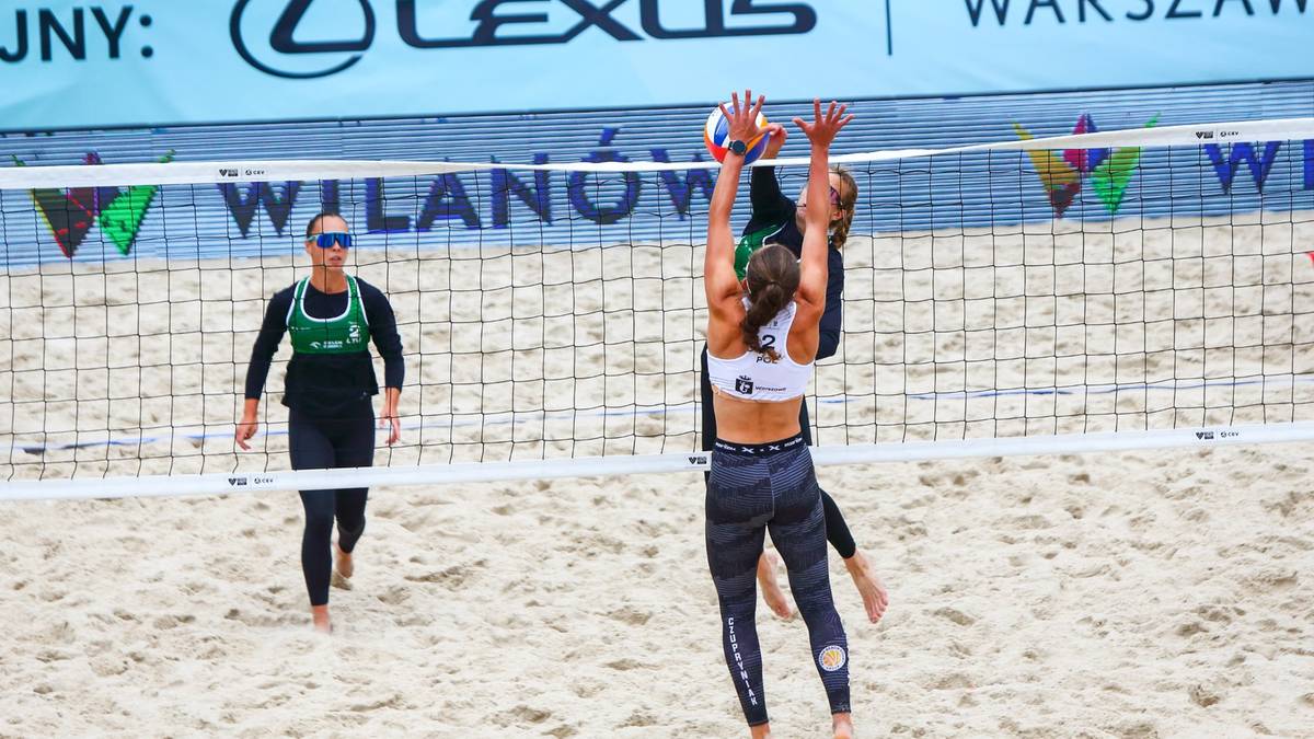 Dwie siatkarki plażowe w akcji przy siatce podczas meczu. Jedna z zawodniczek pr&oacute;buje zablokować atak, druga z drużyny przeciwnej wykonuje wyskok do zagrywki. Na drugim planie widoczny jest sędzia i fragment bandy reklamowej.