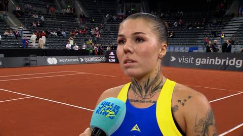 Portret młodej kobiety z tatuażami na szyi i ramionach, trzymającej mikrofon podczas wywiadu na korcie tenisowym.