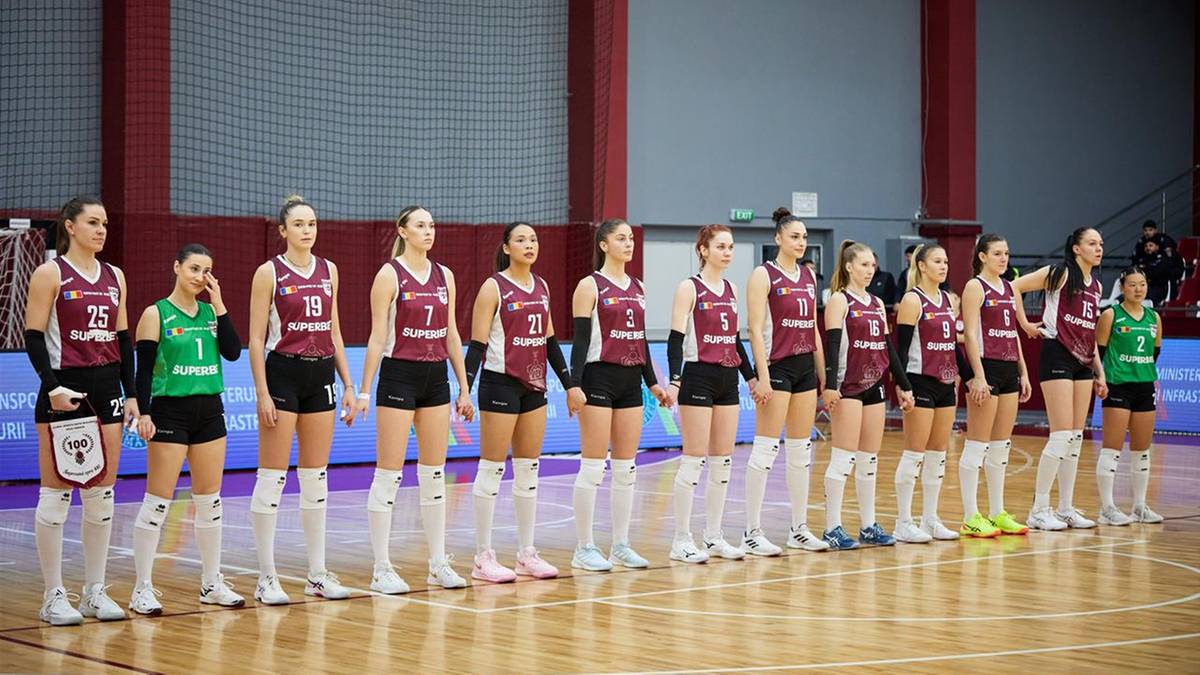 Grupa kobiecej drużyny siatkarskiej stojąca w rzędzie na boisku, w strojach sportowych z numerami na koszulkach.