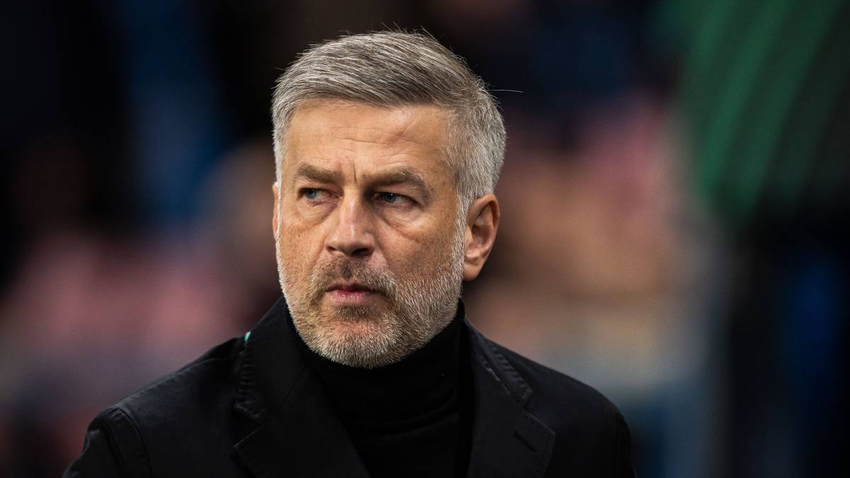 Portret mężczyzny w średnim wieku, o kr&oacute;tkich siwych włosach i zaroście, ubranego w czarny golfik i marynarkę. Jego wyraz twarzy jest poważny.