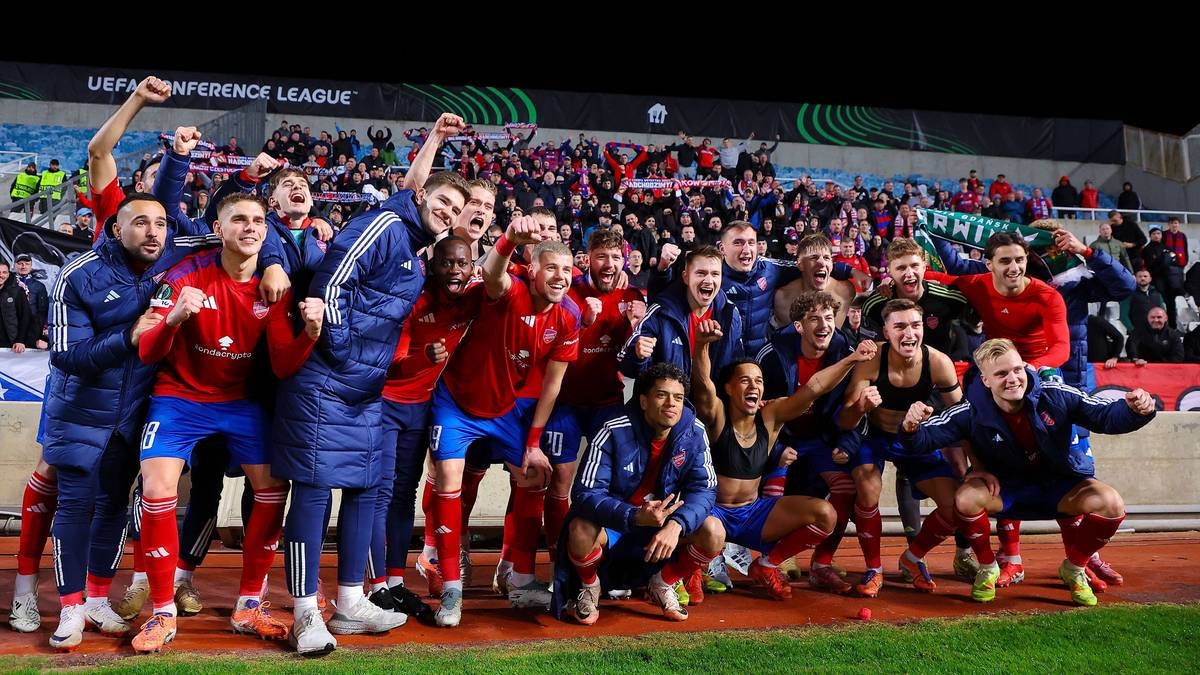 Drużyna piłkarska świętująca zwycięstwo na murawie stadionu, z kibicami na trybunach w tle.