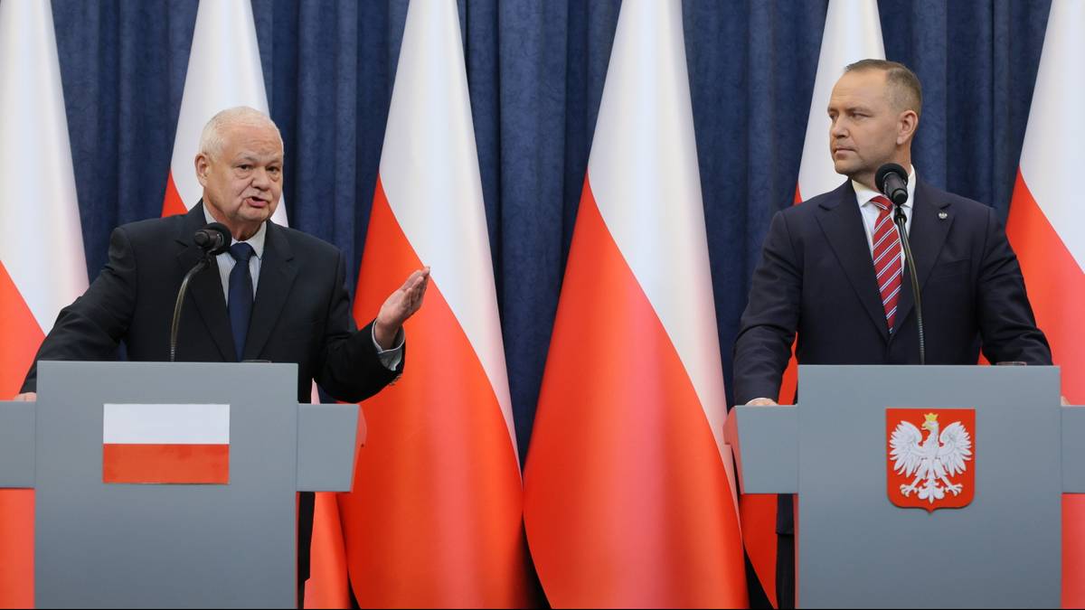 Dw&oacute;ch mężczyzn w garniturach stoi za m&oacute;wnicami, na kt&oacute;rych widnieją polskie flagi i godło.