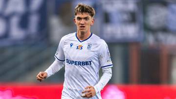 Piłkarz w białym stroju z numerem 27 na plecach prowadzi piłkę po boisku.