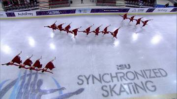 Dwie grupy łyżwiarek synchronicznych w czerwonych strojach wykonują choreografię na lodowisku podczas zawod&oacute;w.