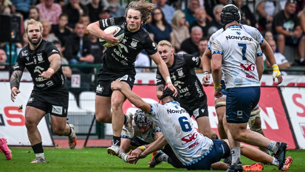 Zawodnicy drużyny rugby w czarnych strojach walczą o piłkę. Jeden z graczy z numerem 5 na koszulce biegnie z piłką w ręku, otoczony przez przeciwników w jasnych strojach. W tle widać trybuny wypełnione kibicami.