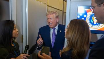 Donald Trump w garniturze i krawacie, otoczony przez dziennikarzy z mikrofonami, przemawia z podniesioną ręką.