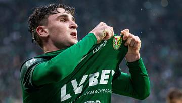 Piłkarz w zielonej koszulce zdejmuje ją z radości, z tyłu widać trenera.