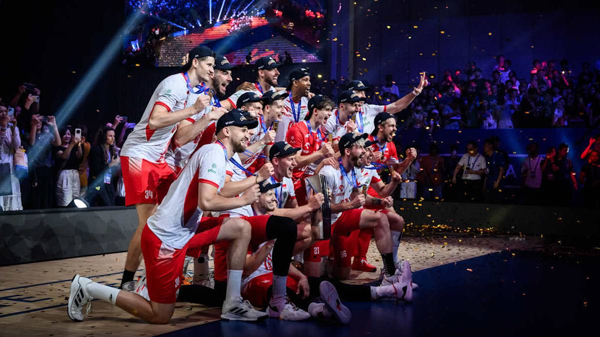 Siatkarze w strojach sportowych świętują zwycięstwo, pozując do zdjęcia w grupie. Niektórzy trzymają medale, inni obejmują się lub unoszą ramiona. Tło stanowi rozmyta widownia i oświetlenie sceniczne.