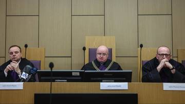 Trzech sędzi&oacute;w w togach sądowych siedzi za stołem w sali sądowej przed herbem Polski.