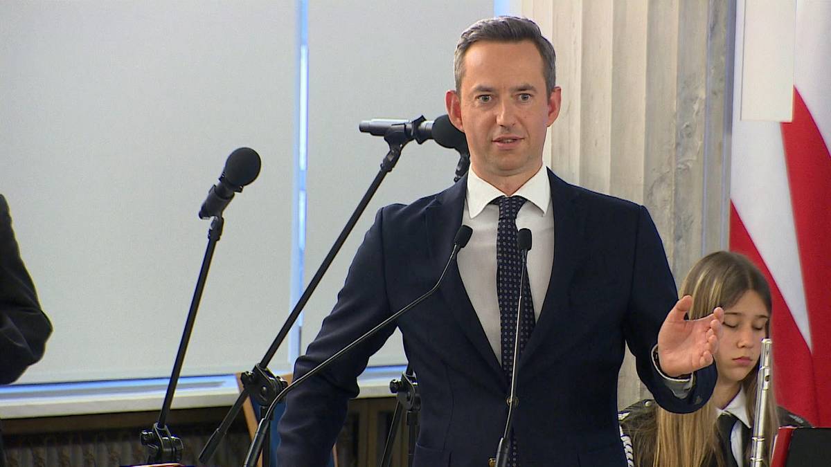 Mężczyzna w granatowym garniturze i białej koszuli z krawatem przemawia przy m&oacute;wnicy. Obok niego stoi dziewczynka.