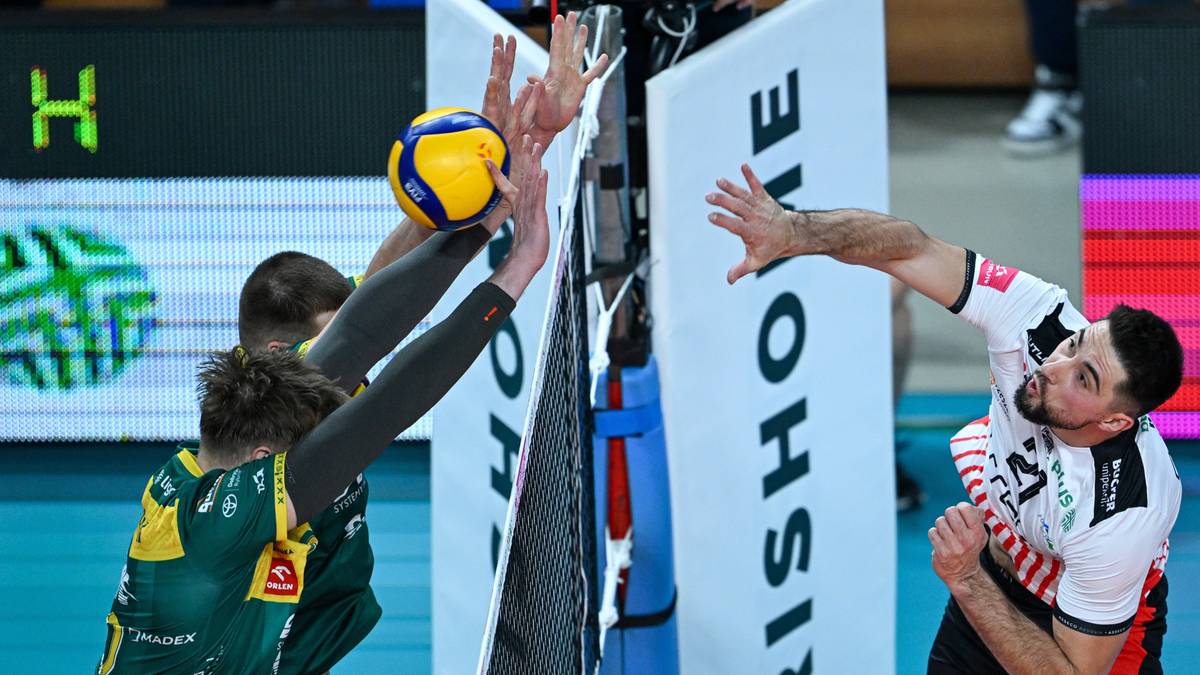 Siatkarz w czarnej koszulce z numerem 21 próbuje zablokować atak przeciwnika w zielonej koszulce. Piłka jest tuż nad siatką.