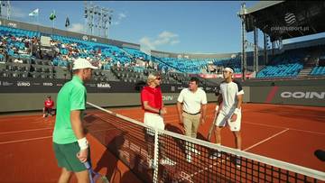 Czterech mężczyzn na korcie tenisowym, dw&oacute;ch w strojach sportowych po przeciwnych stronach siatki, z trybunami pełnymi widz&oacute;w w tle.