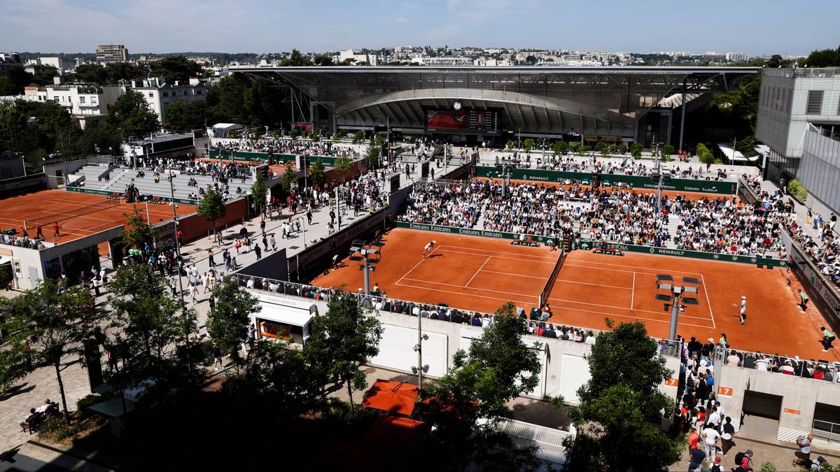 Widok z lotu ptaka na kompleks kort&oacute;w tenisowych podczas meczu. Widać kilka ziemnych kort&oacute;w otoczonych trybunami z publicznością. W tle znajduje się nowoczesny, zadaszony stadion oraz budynki otoczone zielenią.