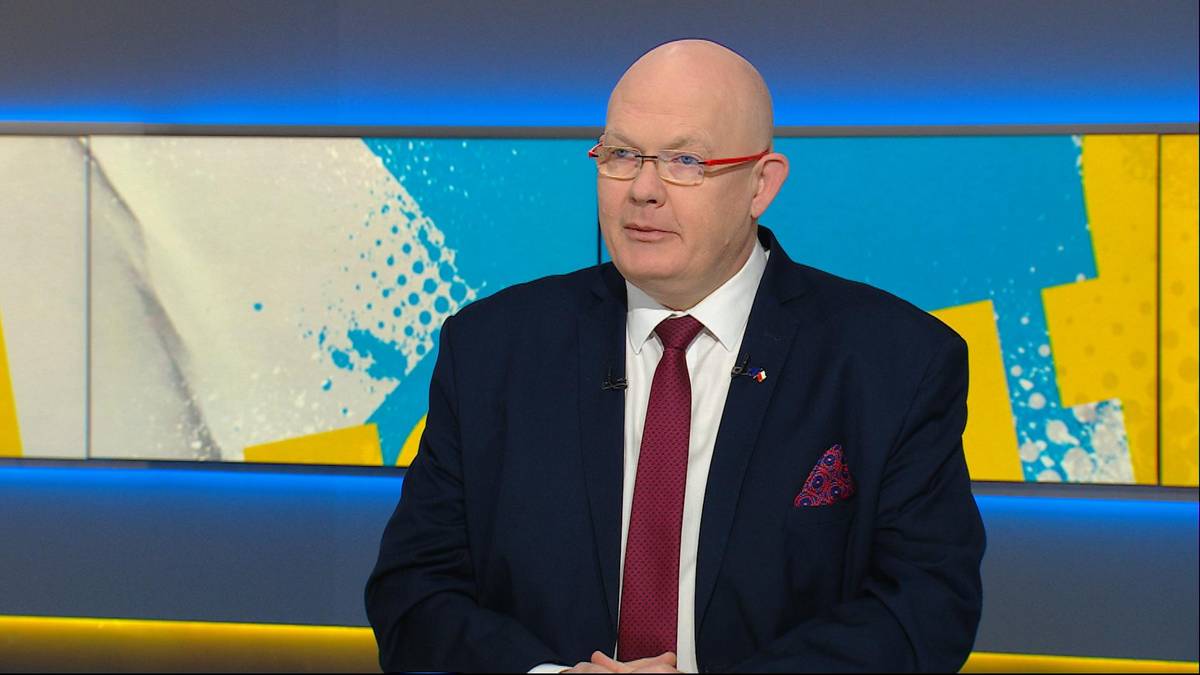 Mężczyzna w garniturze i okularach siedzi przed kolorowym tłem.