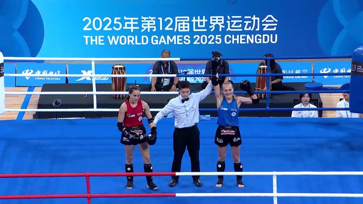 Trzy osoby stoją na niebieskiej macie w ringu do muay thai. W środku stoi mężczyzna w białej koszuli i czarnym krawacie, trzymając za ręce dwie kobiety ubrane w stroje sportowe.