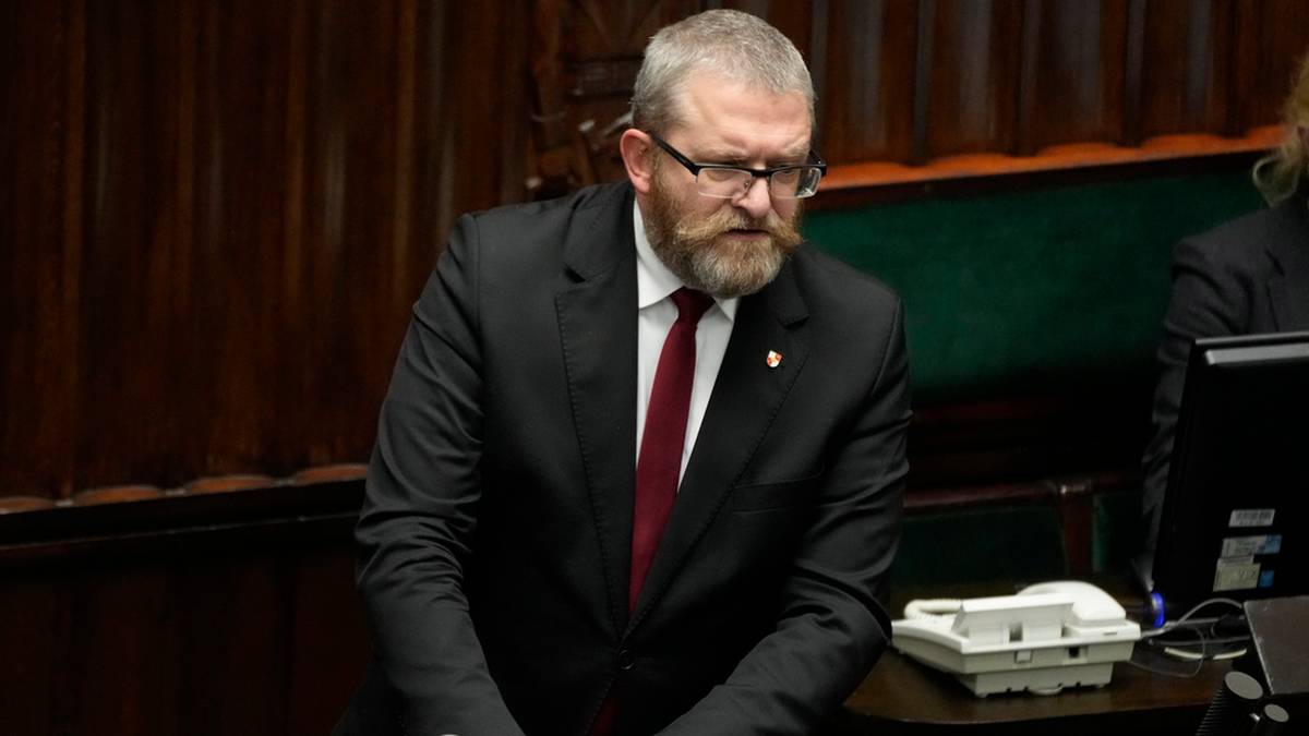 Grzegorz Braun w garniturze i okularach stoi przy mównicy.