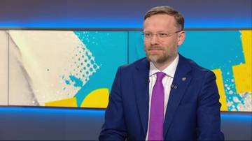 Mężczyzna w granatowym garniturze i fioletowym krawacie, w okularach, siedzi w studiu telewizyjnym.