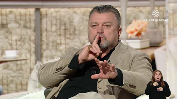 Mężczyzna w średnim wieku, Roman Kosecki, siedzi w studiu telewizyjnym i gestykuluje, wskazując palcem na usta, co sugeruje prośbę o ciszę.