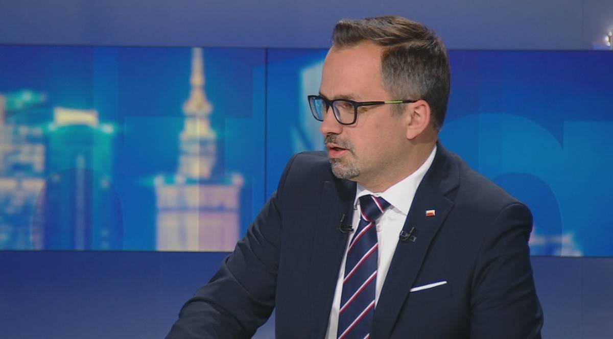 Mężczyzna w garniturze i okularach wypowiadający się w studio telewizyjnym na tle niebieskiej scenografii.