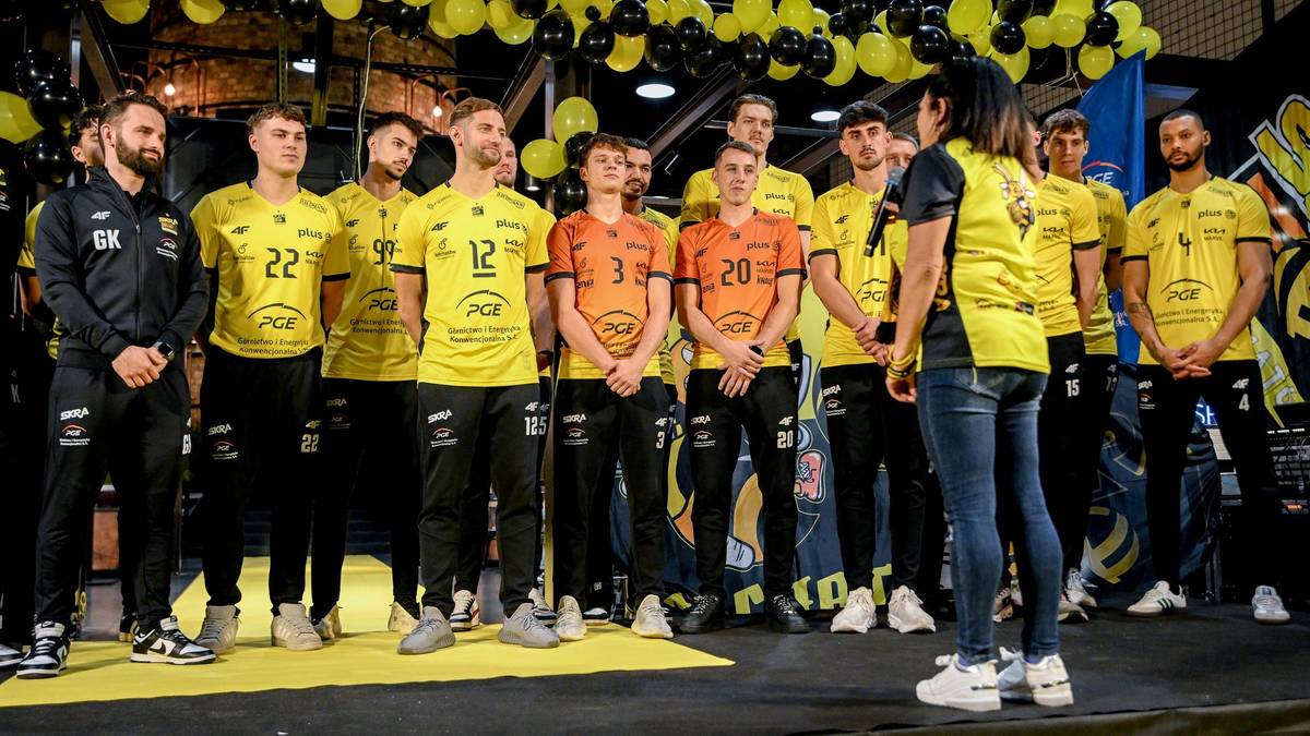 Grupa siatkarzy PGE GiEK Skra Bełchatów w żółtych i czarnych strojach sportowych, stojąca w rzędzie. Kobieta stoi przed nimi, patrząc w ich stronę.
