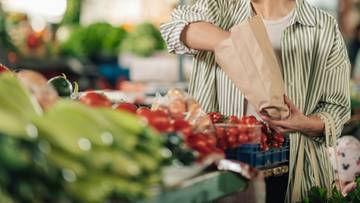 Osoba wybiera pomidory na targu spożywczym, trzymając papierową torbę.