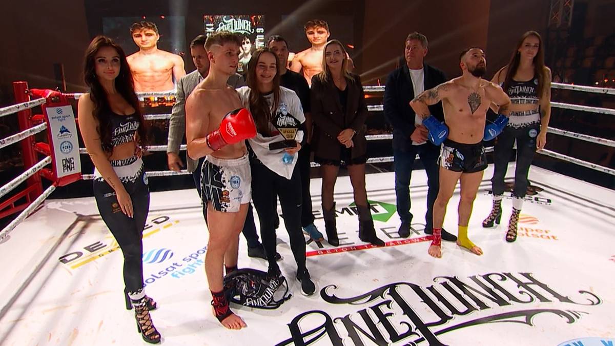Grupa osób, w tym mężczyźni i kobiety, stoi na ringu bokserskim. Jeden z mężczyzn trzyma rękawicę bokserską.