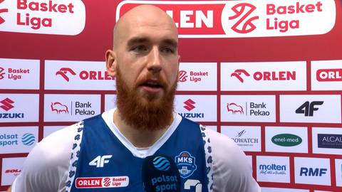 Adrian Bogucki, zawodnik koszyk&oacute;wki z czerwoną brodą, ubrany w niebiesko-białą koszulkę, wypowiada się do mikrofonu.