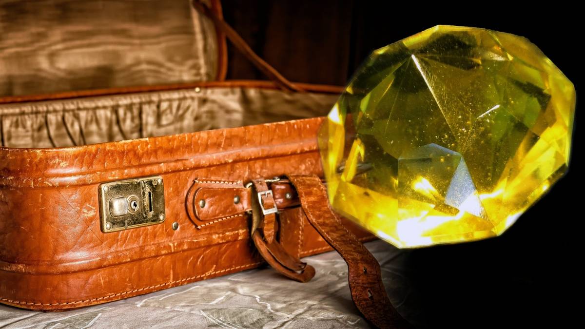 Zbliżenie na otwartą, brązową, sk&oacute;rzaną walizkę z otwartą klapą, obok kt&oacute;rej leży duży, ż&oacute;łty, fasetowany kamień przypominający diament.
