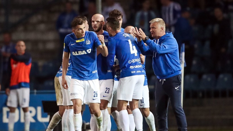 Grupa piłkarzy w niebieskich strojach cieszy się na boisku piłkarskim, a trener w tym samym kolorze bluzy podaje im dłonie.