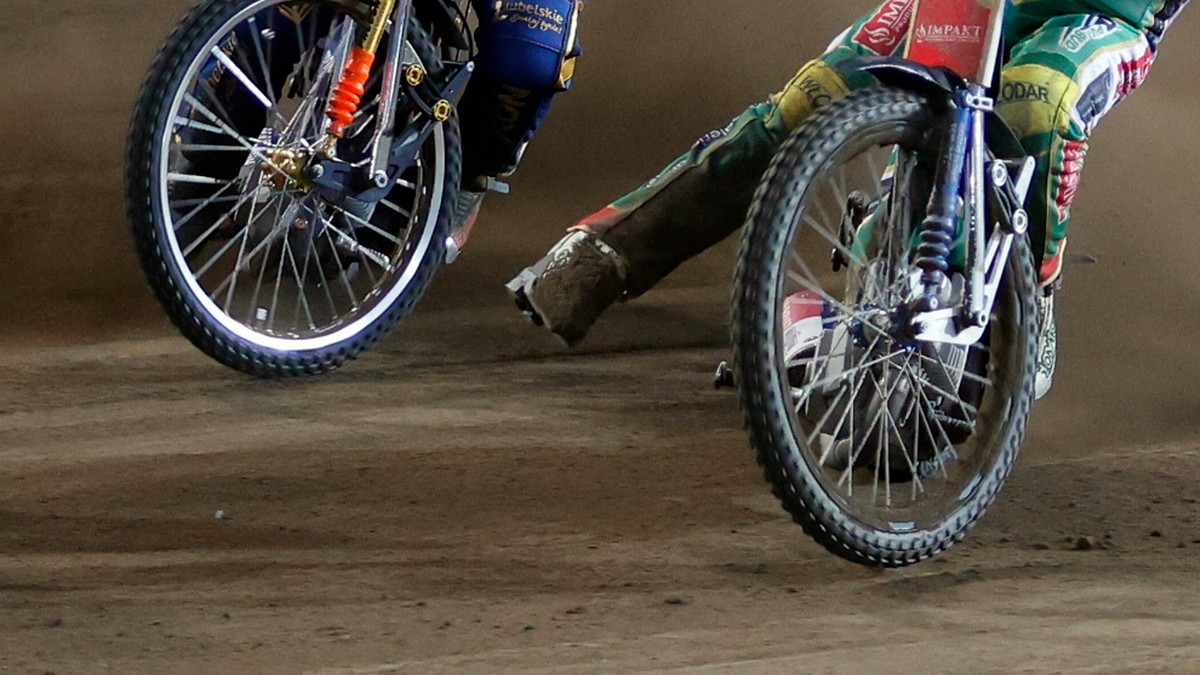 Dwa motocykle żużlowe na torze, widok z bliska na przednie koła i nogi zawodnik&oacute;w.