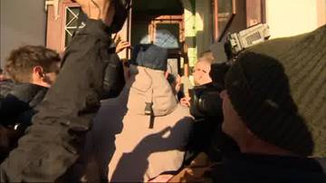 Grupa ludzi, w tym policjanci, w trakcie awantury przed budynkiem. Widoczne kamery.