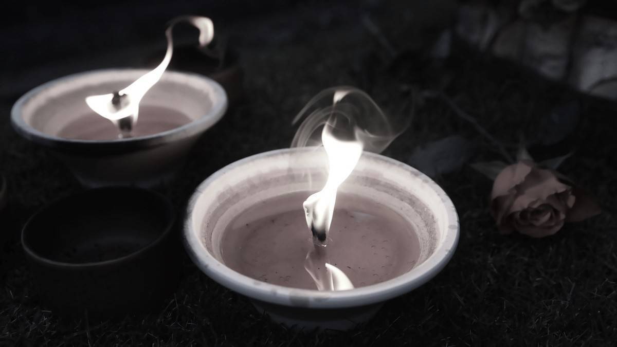 Dwa znicze zapalone na ciemnym tle, obok nich leży pojedyncza r&oacute;ża. Płomienie świec rzucają migotliwe światło.