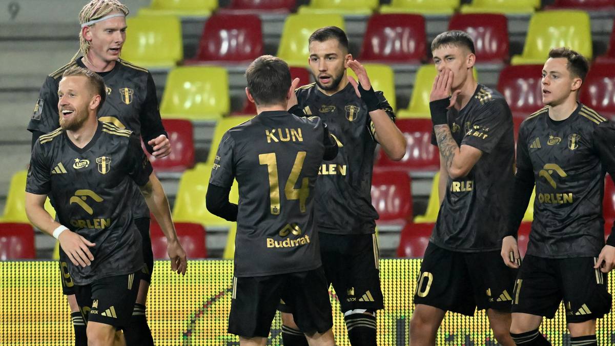 Piłkarze w czarnych strojach z żółtymi elementami stoją blisko siebie na boisku, niektórzy rozmawiają, inni patrzą w kierunku kamery.