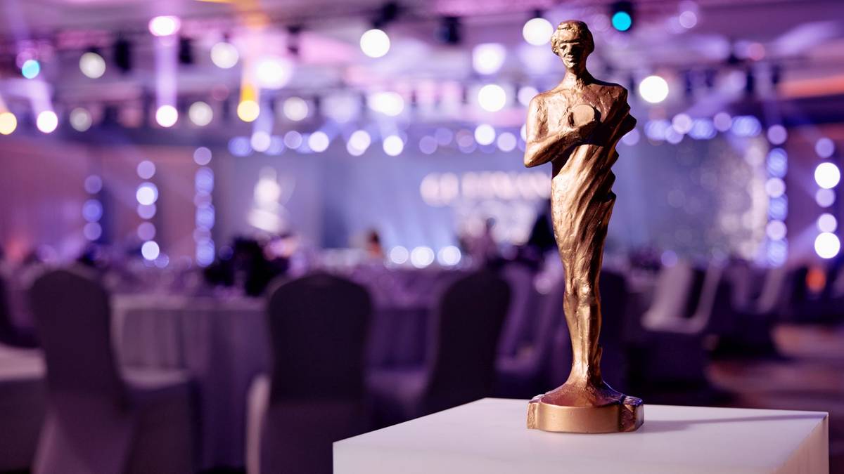 Złota statuetka nagrody plebiscytu "Guttmanny" na białym postumencie, z rozmytym tłem sali bankietowej.