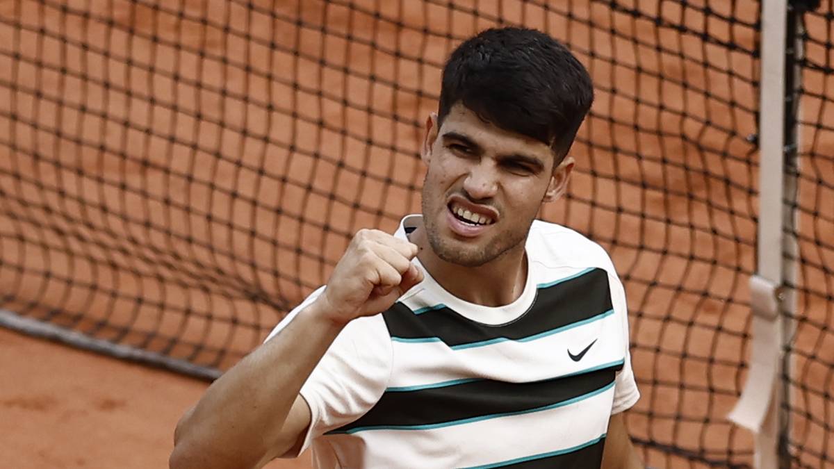 Carlos Alcaraz, uśmiechnięty, świętujący zwycięstwo podczas meczu tenisa na korcie ziemnym. Ma na sobie białą koszulkę w czarne paski i unosi zaciśniętą pięść w geście radości. W tle widoczna jest siatka tenisowa.