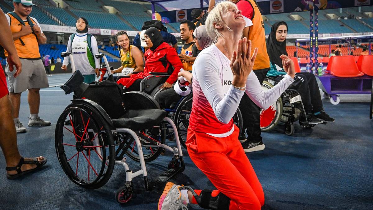 Kobieta w czerwonym stroju sportowym klęczy na niebieskiej nawierzchni, unosząc ręce w geście radości. Za nią widoczni są inni ludzie i wózki inwalidzkie.