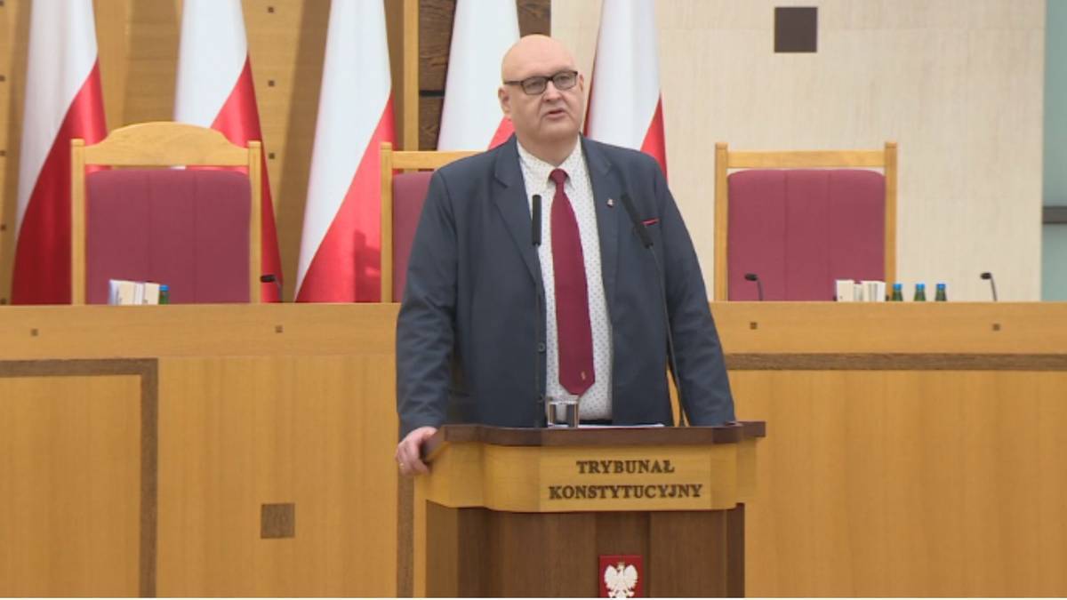 Mężczyzna w garniturze i krawacie stoi za m&oacute;wnicą z napisem "Trybunał Konstytucyjny" i przemawia.