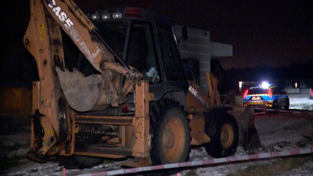 Koparko-ładowarka na placu budowy w nocy, w tle policyjne światła.