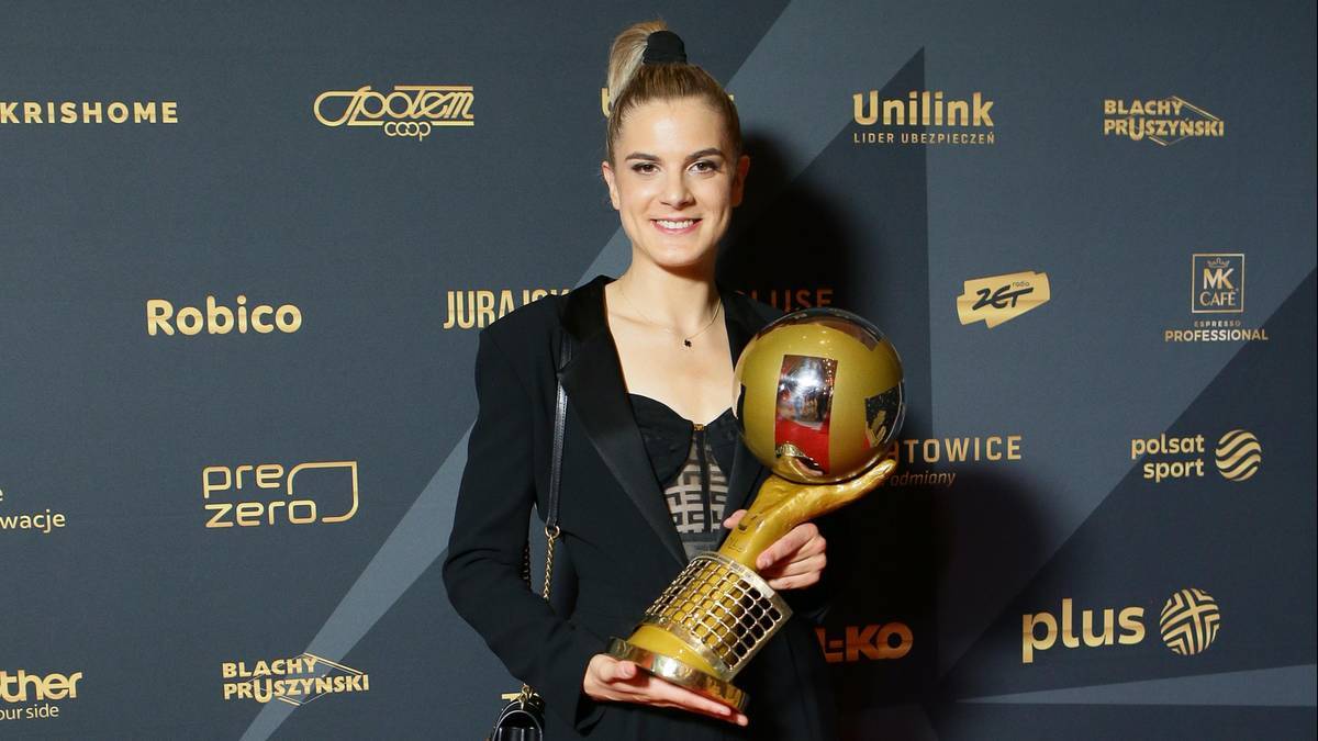 Uśmiechnięta siatkarka trzyma złote trofeum na tle ściany z logo sponsor&oacute;w.