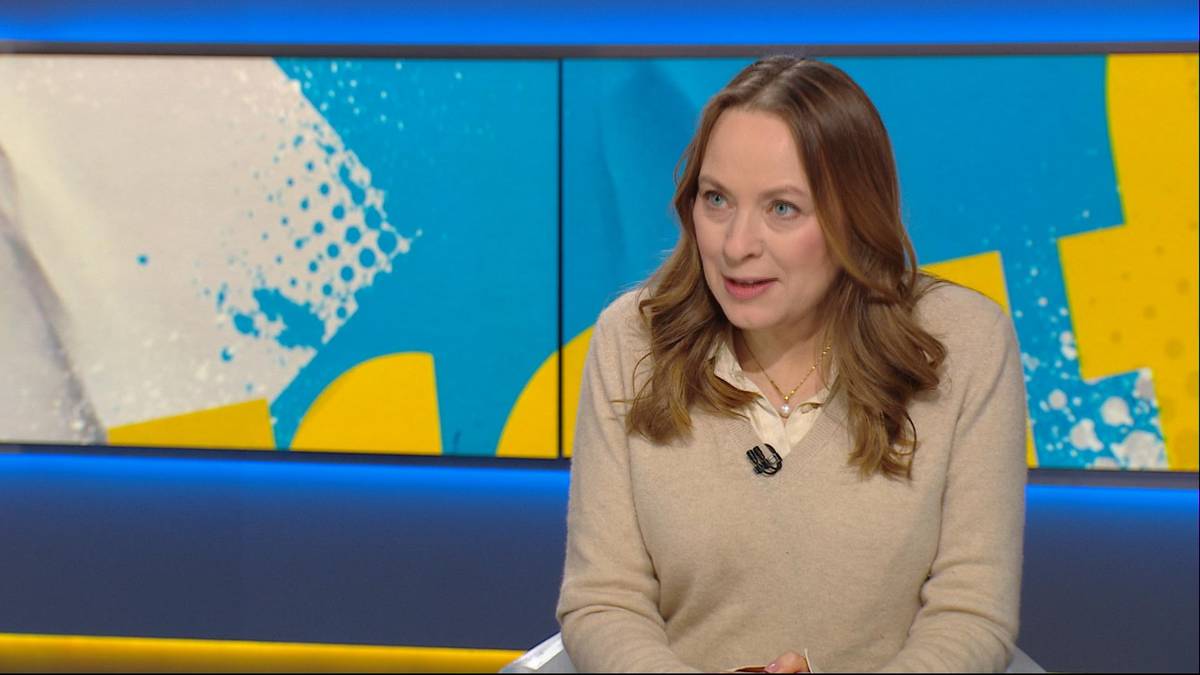 Kobieta w beżowym swetrze siedzi w studiu telewizyjnym, z jasnobrązowymi włosami i naszyjnikiem na szyi. Tło stanowi kolorowa grafika.