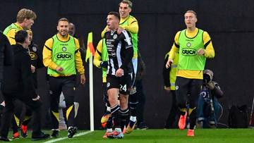 Piłkarz w czarno-białej koszulce, numer 24, cieszy się z zdobytej bramki, otoczony przez kolegów z drużyny w żółtych kamizelkach.