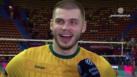 Młody mężczyzna, prawdopodobnie siatkarz, w ż&oacute;łtej koszulce z zielonym kołnierzykiem, uśmiecha się szeroko do kamery. Trzyma w ręku mikrofon z logo Polsat Sport.