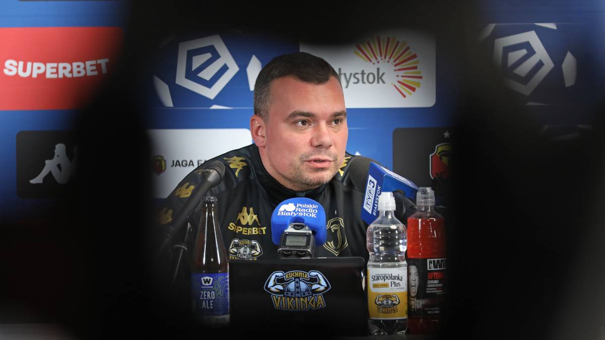 Adrian Siemieniec, trener Jagiellonii Białystok, siedzi przy stole podczas konferencji prasowej z mikrofonem przed nim.