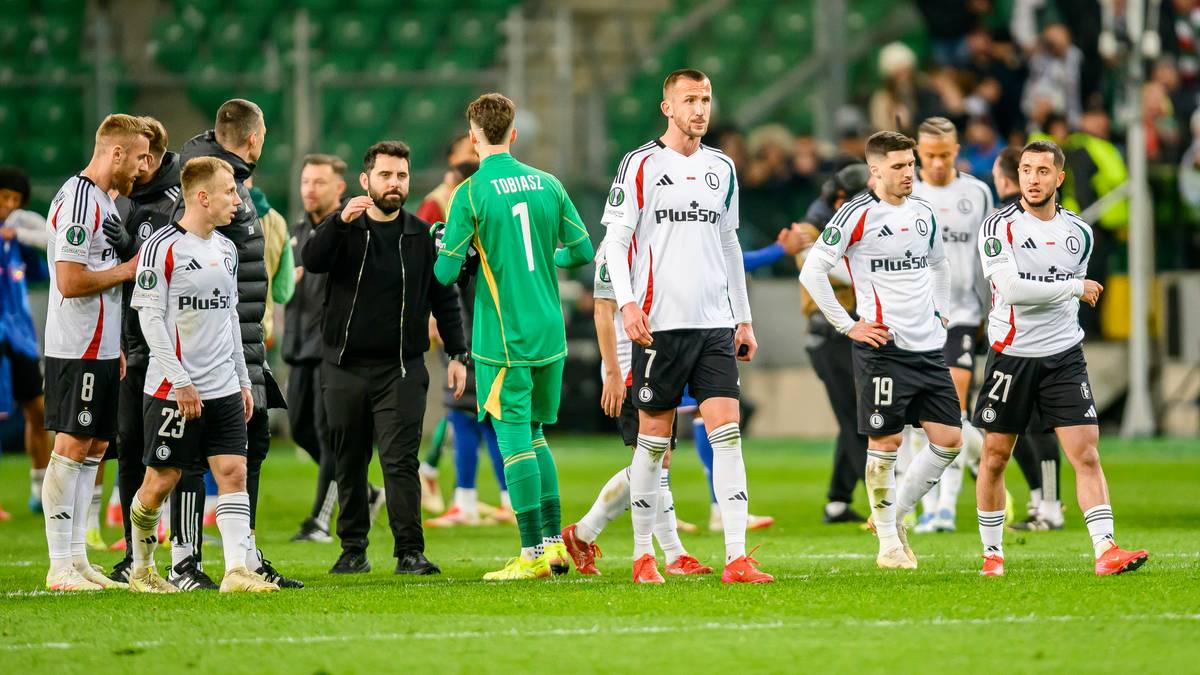 Grupa piłkarzy w strojach Legii Warszawa stoi na boisku piłkarskim.