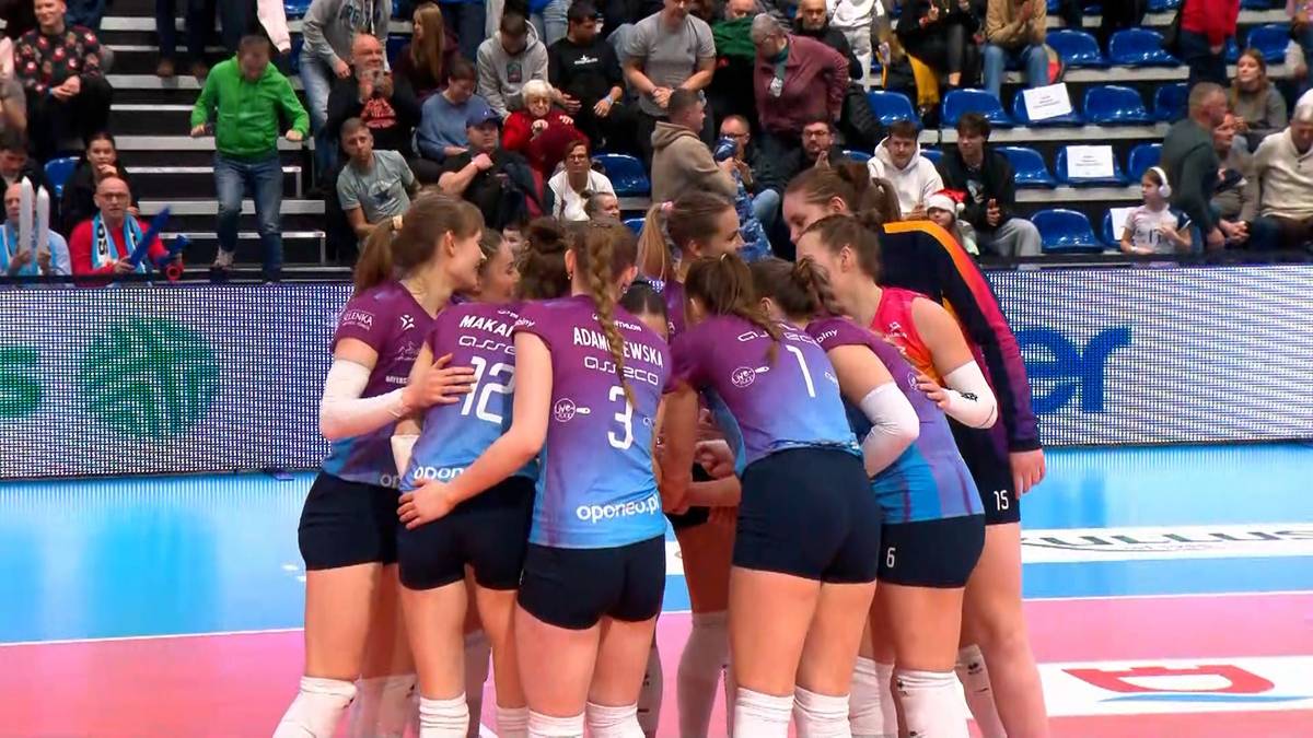 Grupa siatkarek w kolorowych strojach sportowych zbiera się w geście jedności na boisku.