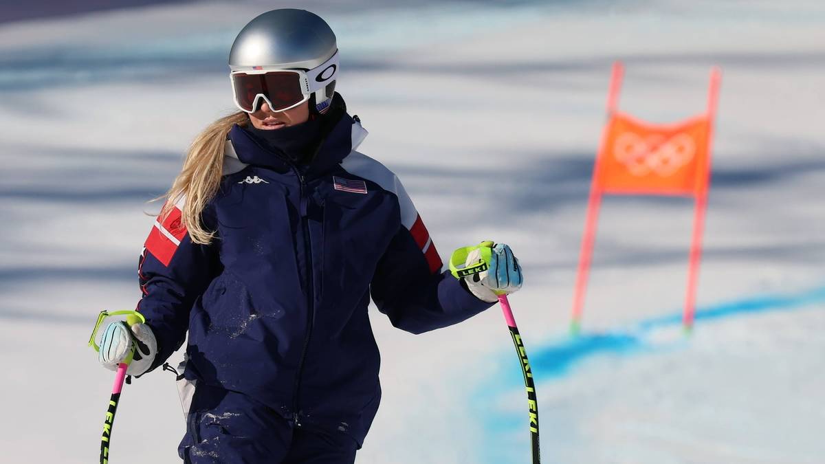 Narciarka alpejska w kasku i goglach narciarskich, ubrana w kombinezon, z kijkami narciarskimi, na tle stoku narciarskiego z tyczkami.