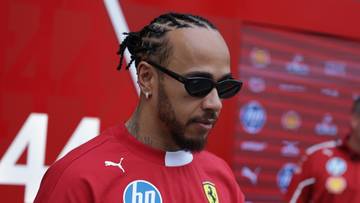 Lewis Hamilton w czerwonym kombinezonie wyścigowym z logotypami sponsorskimi, noszący okulary przeciwsłoneczne.