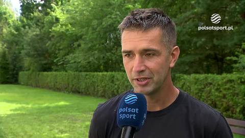 Portret Piotra Kobiereckiego, mężczyzny w średnim wieku, m&oacute;wiącego do mikrofonu z logo "Polsat Sport". W tle zielona trawa, żywopłot i drzewa.