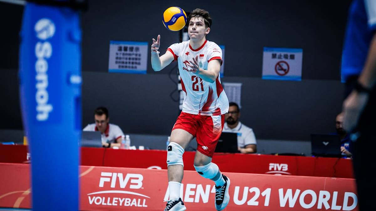 Młody mężczyzna w stroju siatkarskim (biała koszulka z numerem 21, czerwone spodenki) wykonuje zagrywkę podczas meczu siatk&oacute;wki.